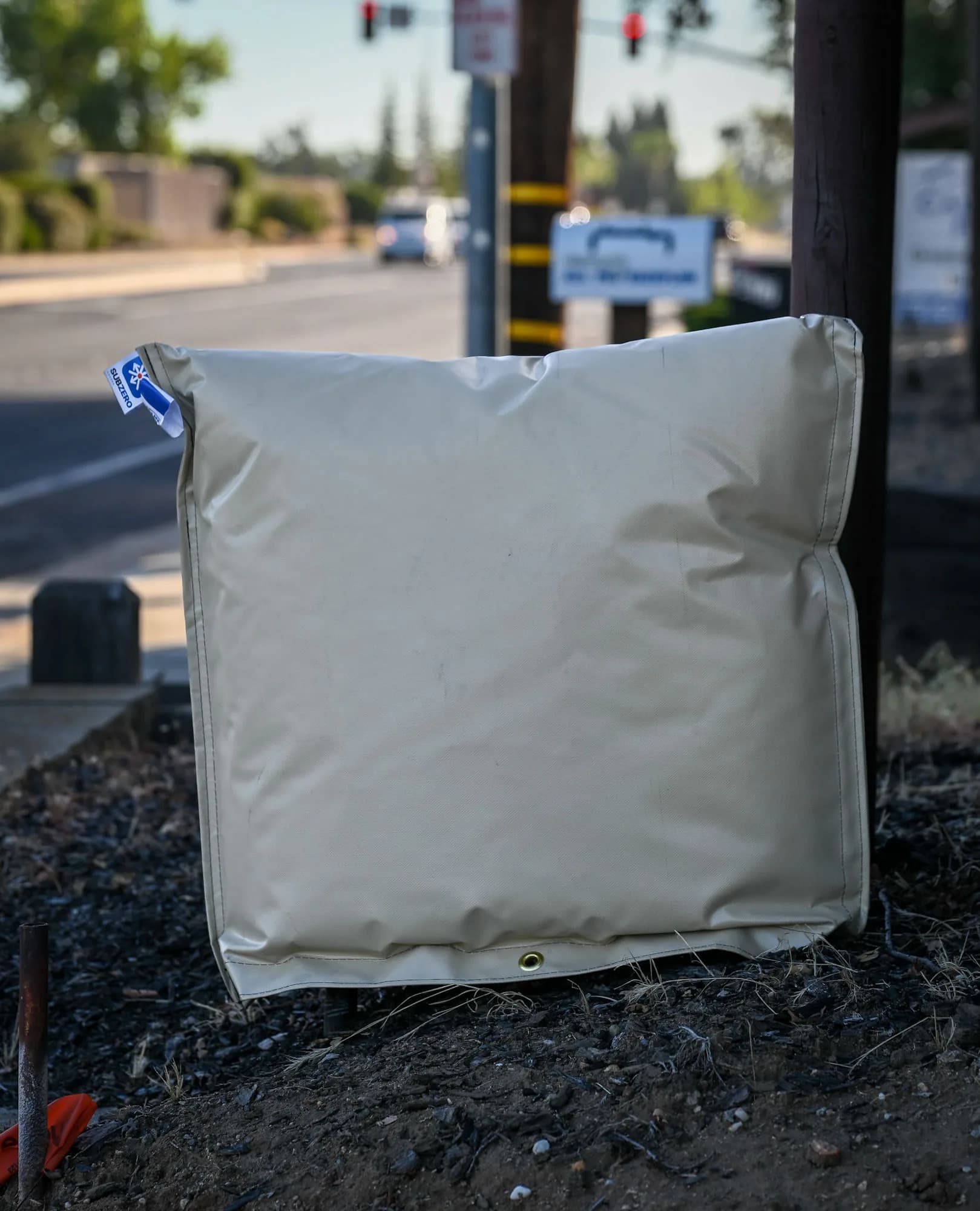 Small tan insulated backflow cover on a roadside irrigation assembly at golden hour, with the Sub Zero Covers product tag visible at the top seam.